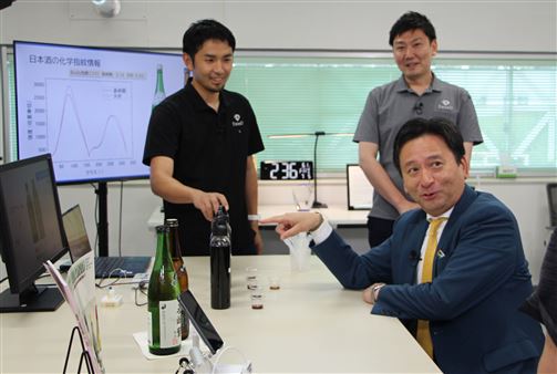 (3)コーヒーの飲み比べを行いAIソムリエの解析結果と照らし合わせる山口知事を、囲むようにして立って見るエクステンドの皆さんの様子。