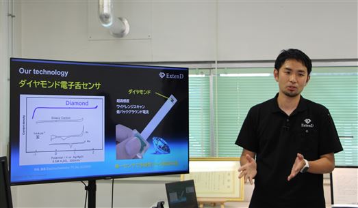 (2)モニターの横に立ち技術について説明する、エクステンドCEO（最高技術責任者）の大曲氏.